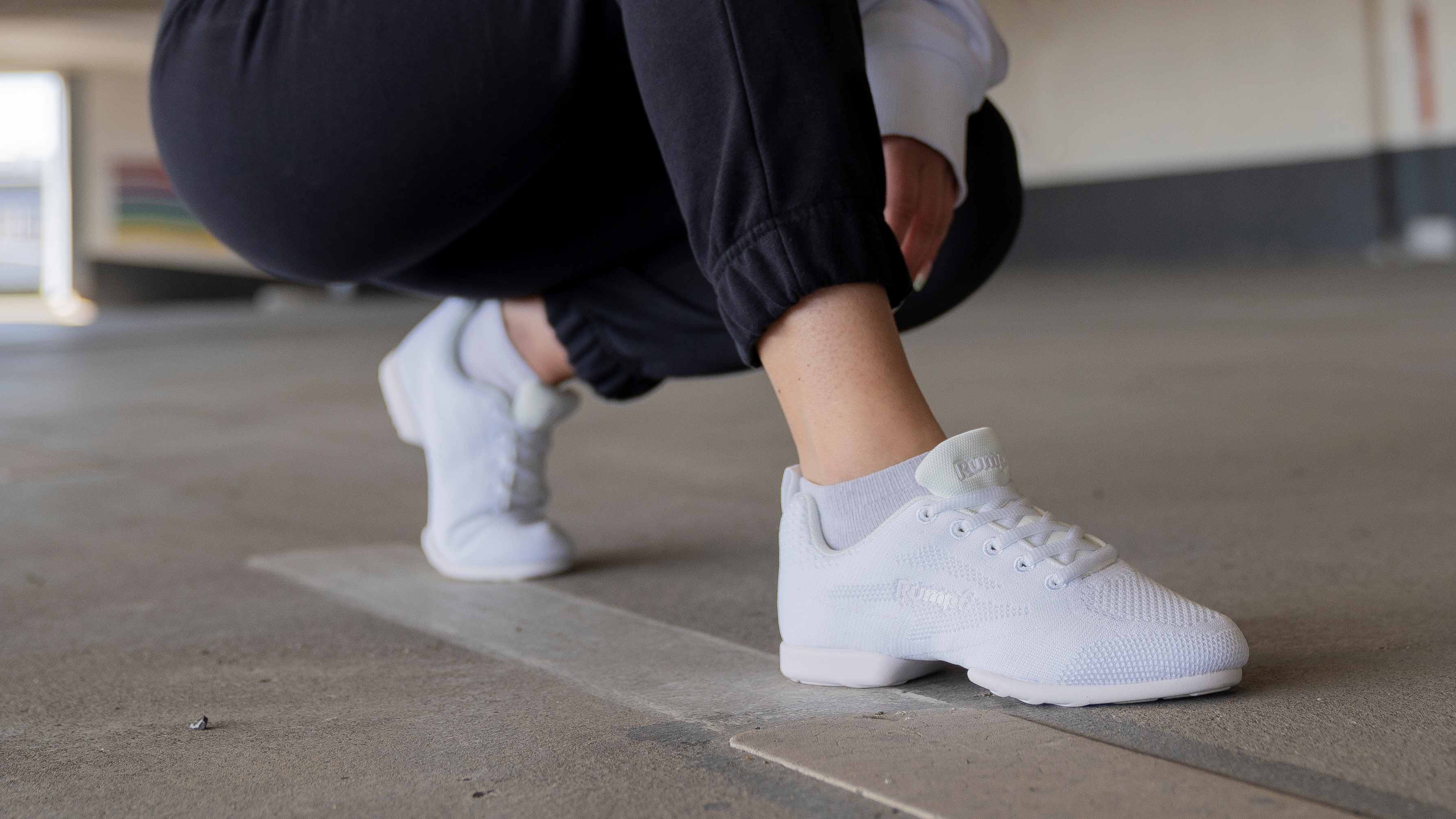 Hockende Person in weißen Turnschuhen und schwarzen Hosen in einer industriellen Umgebung.